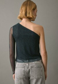 Asymmetrisches Oberteil mit einem dunklen Blumenmuster, einem durchsichtigen langen Ärmel und einem einfarbigen Stoff auf der gegenüberliegenden Seite, kombiniert mit hoch taillierten Jeans.
