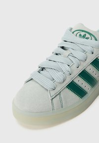 Gaiši pelēkas zamšādas sporta kurpes ar biezām gaiši zilām auklām, zaļām ādas svītrām un zaļu logotipu uz mēles, uz balta fona.