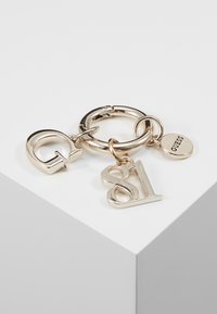 Sleutelhanger in goudkleur met drie bedels: een gestileerde 'G', '81' en een ronde hanger met de inscriptie "GUESS." Voorzien van een gestructureerde ring en een gepolijste afwerking.