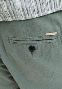 Πίσω τσέπη πράσινου υφάσματος με μαύρο κουμπί και μικρή ταμπέλα σε ταμπά χρώμα που γράφει "Jack & Jones" κάτω από το ρεβέρ ενός λευκού και γκρι ριγωτού πουκαμίσου.