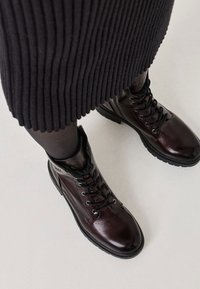 Burgunderfarbene Schnürstiefeletten mit glänzender Textur, schwarzen Akzenten und robuster Gummisohle, die über einem gerippten schwarzen Rock platziert sind.
