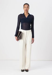 Navy, fitted, long-sleeve knit top with a collar. Paired with cream, high-waisted, wide-leg trousers. Holding a brown clutch. Black heels.