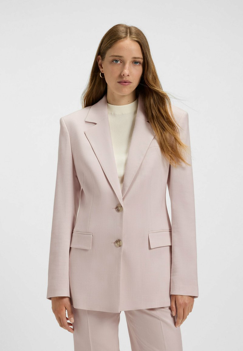 Helle rosa Blazer mit spitzen Revers, zwei Knöpfen, Fronttaschen und taillierter Form, aus glattem Stoff gefertigt. Kombiniert mit einem leichten Pullover.