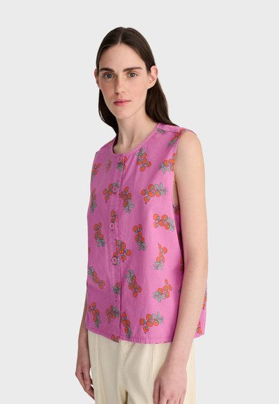 Jeune femme aux longs cheveux noirs portant un chemisier sans manches rose à motif de cerises orange et un pantalon beige, debout devant un fond clair.