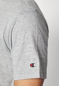 Graues T-Shirt mit kurzen Ärmeln aus weichem Stoff, das ein kleines gesticktes Logo auf dem linken Ärmel trägt. Lockerer Schnitt mit glatter Textur.
