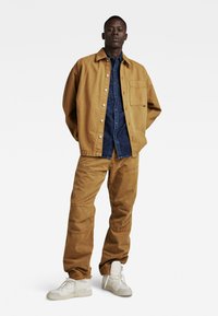 Beige katoenen jack, blauwe denim overhemd eronder en beige katoenen broek. Witte sneakers maken de outfit compleet, met een ontspannen pasvorm en zakken.