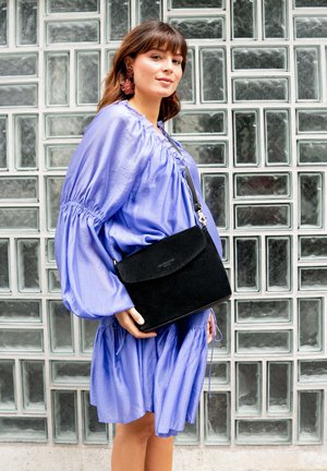Woman in a loose lilac dress with puffed sleeves holds a black suede crossbody bag against a glass block wall background.