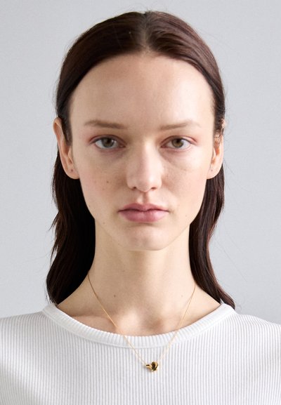 Gold necklace with a small, abstract pendant worn over a white ribbed shirt. The background is a neutral gray. Hair is dark and straight.