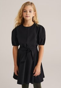 Crna haljina kratkih rukava s okruglim izrezom, strukirana i s mašnom. Suknja se blago širi. Glatka tkanina s klasičnim dizajnom.