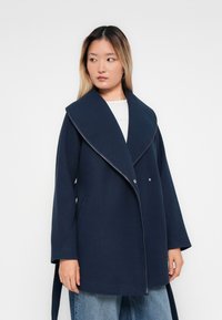 Femme portant un manteau oversized bleu marine avec de larges revers, assorti à un jean bleu clair et un haut blanc, regardant sur le côté.
