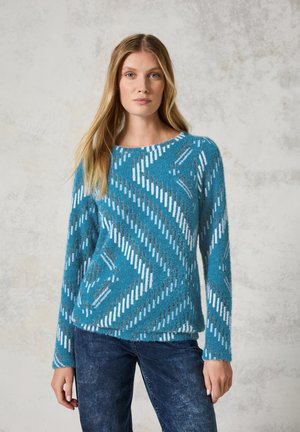 Turkis sweater med et zigzag mønster i hvid og grå. Blød, fuzzy tekstur og lange ærmer. Rund halsudskæring, parret med mørke jeans.