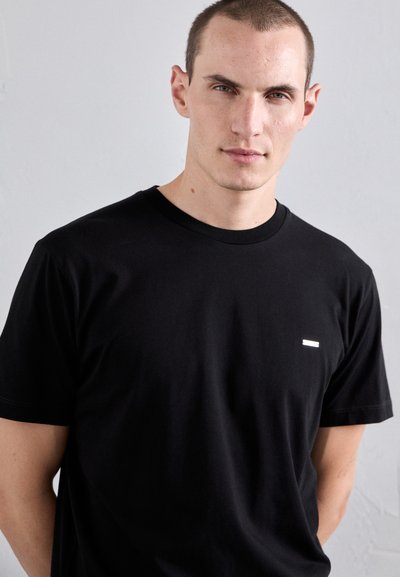 T-shirt nera in cotone con collo a giro e maniche corte, caratterizzata da un piccolo accento rettangolare bianco sul petto. Tessuto liscio.