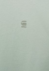 T-shirt vert clair en tissu doux, arborant un logo subtil "RAW" dans une teinte légèrement plus foncée, positionné sur la poitrine.