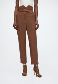 Γυναίκα φορεί υψηλόμεσα καφέ παντελόνια με μοναδική ζώνη σε σχήμα V με κουμπιά και χρυσά πέδιλα με λεπτά λουριά και ψηλό τακούνι.