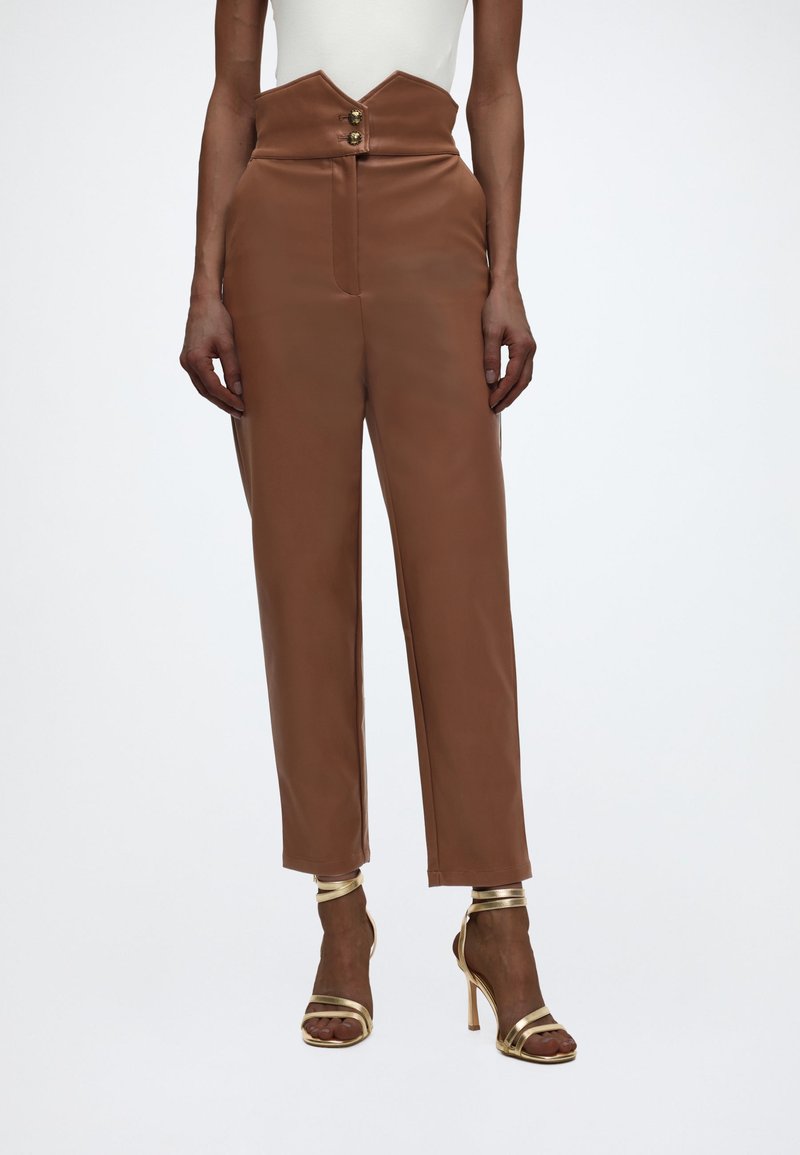 Γυναίκα φορεί υψηλόμεσα καφέ παντελόνια με μοναδική ζώνη σε σχήμα V με κουμπιά και χρυσά πέδιλα με λεπτά λουριά και ψηλό τακούνι.