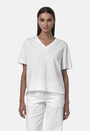 Donna con capelli lisci e scuri che indossa una maglietta bianca a collo a V e pantaloni bianchi, in piedi davanti a uno sfondo chiaro e semplice.