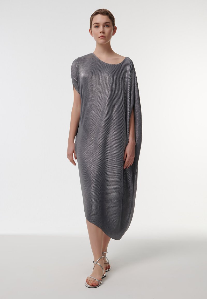 Vestido metálico gris, de largo hasta la rodilla, con diseño asimétrico, mangas anchas y textura suave. Combinado con sandalias plateadas de tiras.