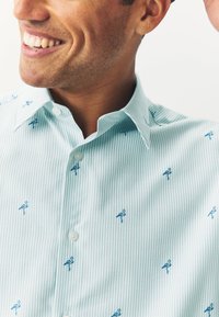 Camicia a righe azzurro chiaro con ricamo di piccoli fenicotteri blu, colletto classico e chiusura con bottoni, realizzata in un misto cotone traspirante.