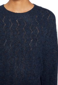 Pull en maille bleu marine avec un motif ondulé et une texture en maille ajourée. Col rond avec bord-côte au niveau de l'encolure et des manches.