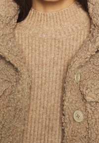 Beiger Pullover mit gerippter Struktur und hohem Kragen, geschichtet unter einem flauschigen beigen Mantel mit großen Knöpfen und weichem, zotteligem Außenmaterial.