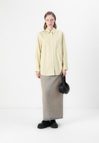 Camicia gialla a righe con bottoni, gonna in denim grigia, scarpe nere basse, tiene in mano una piccola borsa nera. Taglio semplice e rilassato con una texture liscia.