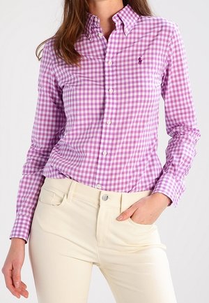 Mujer con camisa de cuadros morados y blancos abotonada y pantalones beige claros, con una mano en el bolsillo y cabello largo y castaño visible.