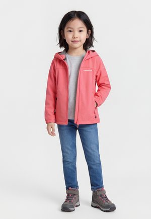 Νεαρή κοπέλα που φοράει κοραλλί μπουφάν με κουκούλα, γκρι μπλούζα, μπλε τζιν και γκρι ορειβατικά μποτάκια, στέκεται με το ένα χέρι στην τσέπη του μπουφάν.