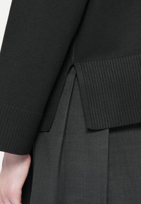 Svart genser med ribbestruktur, som har en splittet og lagdelt design i kanten. Myk tekstur, variabel stofftykkelse.