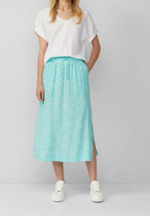 Vrouw in een witte top met opgerolde mouwen en V-hals, turkooise midi-skirt met een patroon en een zijsplit, en witte sneakers, staand tegen een effen achtergrond.