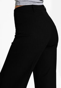 Pantalons noirs avec une texture lisse, taille ajustée et jambes larges et fluides. Le tissu semble extensible et léger.