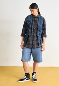 Denimweste mit mehreren Taschen über einem schwarz-grau karierten Hemd, kombiniert mit hellen Jeansshorts und schwarzen High-Top-Sneakers.