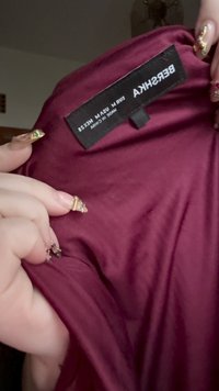 Detailní záběr na bordó látku Bershka s hladkou texturou a černou visačkou, na které jsou uvedeny velikost a pokyny k péči. Je vidět ruka držící látku.