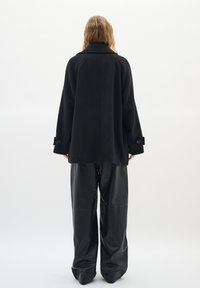 Black wool coat with a wide silhouette, large collar, and buttoned cuffs. Paired with loose-fitting black leather pants.