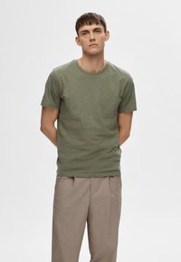 T-shirt vert à manches courtes en tissu doux, avec un col rond, une coupe décontractée et une texture subtile. Porté avec un pantalon beige plissé.