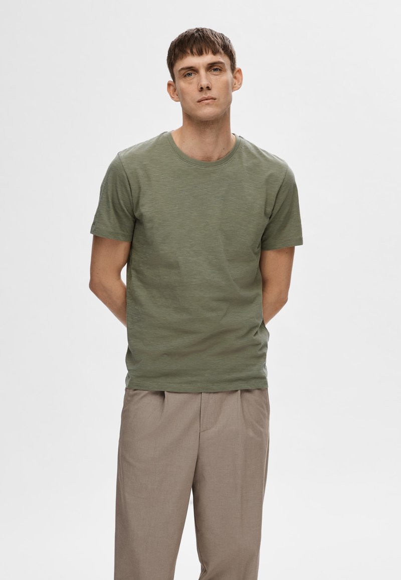 T-shirt vert à manches courtes en tissu doux, avec un col rond, une coupe décontractée et une texture subtile. Porté avec un pantalon beige plissé.