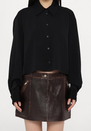 Femme portant un chemisier noir à manches longues boutonné, rentré dans une mini-jupe en cuir marron foncé avec des poches zippées à l'avant.