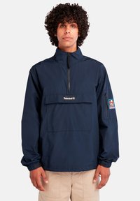 Tamsiai mėlynas, lengvas pūkinis džemperis su pusiau užtrauktuku priekyje, elastiniais rankogaliais ir krūtine kišene, kurioje yra Timberland logotipas ir pleistras.