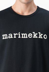 Juodas marškinėlis su apvaliu iškirpimu, pagamintas iš medvilnės, su dideliu baltu tekstu "marimekko", atspausdintu per krūtinę. Lygi tekstūra.