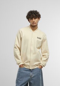 Krémový, zapínací cardigan s rebrovaným golierom, postrannými vreckami a čiernym nápisom "LOS ANGELES" na hrudi. Mäkká, štruktúrovaná tkanina.