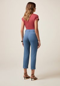 Donna con pantaloni blu arrotolati, maglietta corta color ruggine, braccialetto nero e sandali con tacco a strato in stampa serpente, in piedi con la schiena rivolta.