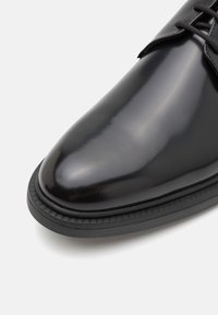 Chaussure habillée en cuir noir brillant avec bout arrondi, semelle noire et lacets noirs sur fond blanc.