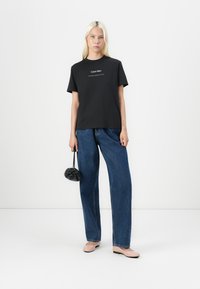 Czarna bawełniana koszulka z białym logo "Calvin Klein", zestawiona z luźnymi niebieskimi dżinsami i jasnoróżowymi balerinami, trzymająca małą czarną torebkę.