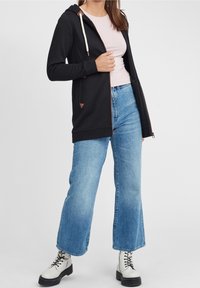 Černá zipová mikina s krémovými šňůrkami, hnědými akcenty, nošená přes růžové tričko a modré široké džíny, doplněná bílými šněrovacími botami.