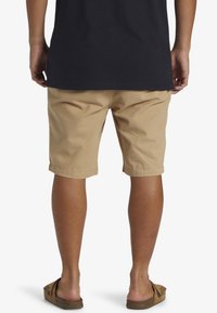 Pantalones cortos de algodón color beige, hasta la rodilla y con un frente plano. Combinando con una camisa oscura, sandalias visibles con una tira texturizada sobre los pies descalzos.