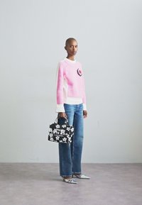 Pull en tricot rose avec des bords blancs et un patch floral, associé à un jean bleu. Tient un sac tote noir avec des motifs de fleurs blanches. Chaussures argentées.