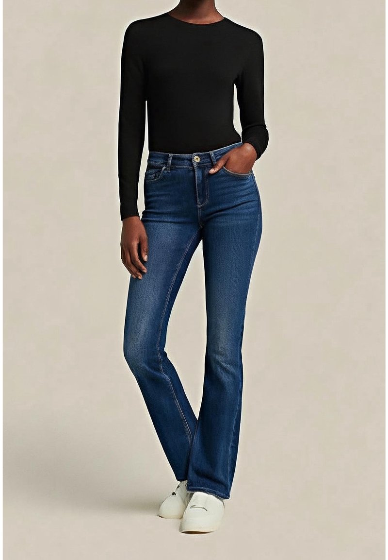 Personne portant un jean droit bleu marine, un haut à manches longues noir et des baskets blanches, debout avec une main dans la poche.