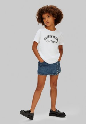 Barn med krøllet hår iført hvid Calvin Klein t-shirt, denimskørt og sorte loafers står med hænderne i lommerne.