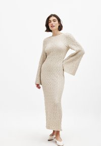 Kootud pika varrukaga kleit heledas beežis, tekstuurse viimistlusega ja laiendatud varrukatega, keha lähedane siluett kuni pahkluuni.