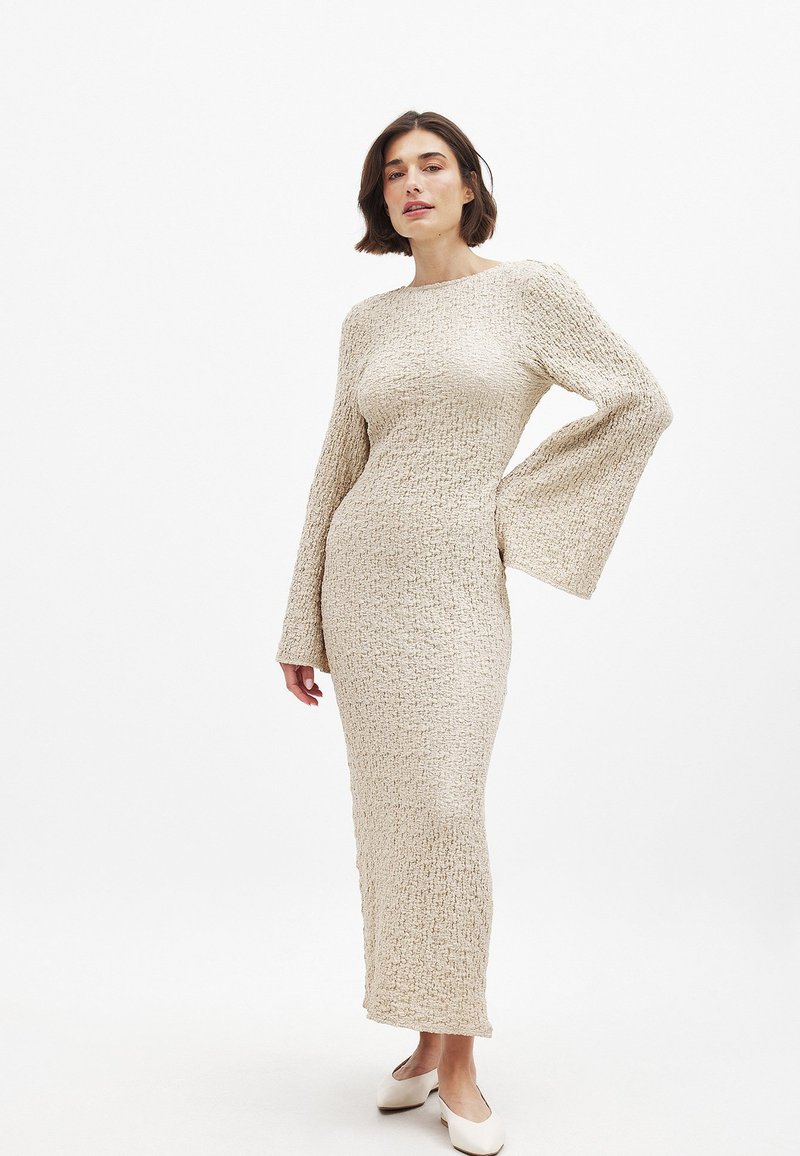 Kootud pika varrukaga kleit heledas beežis, tekstuurse viimistlusega ja laiendatud varrukatega, keha lähedane siluett kuni pahkluuni.