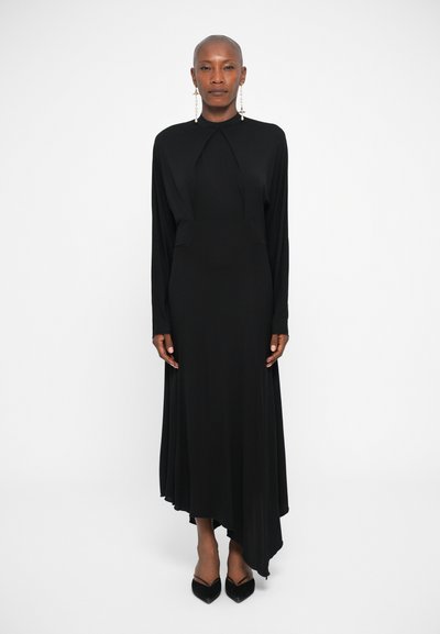 Vestido negro de manga larga con dobladillo asimétrico, tela suave, cuello alto y detalles sutiles de drapeado. Combinado con zapatos negros puntiagudos.